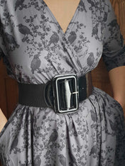 Gray dress with black ravens & blossoms pattern and black belt worn by a person against a wooden door background