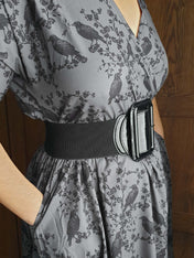 Gray dress with black ravens & blossoms pattern and black belt worn by a person against a wooden door background