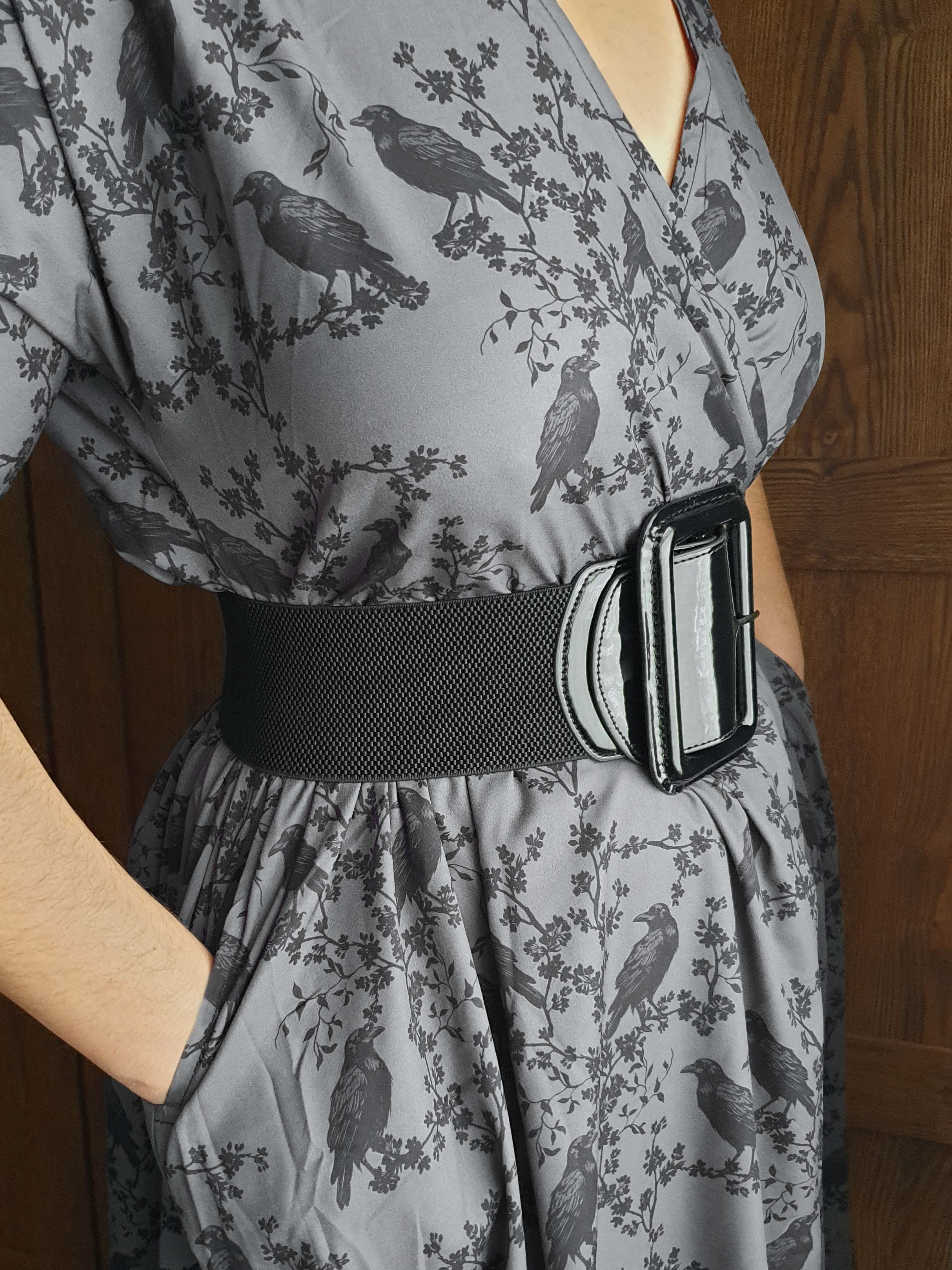 Gray dress with black ravens & blossoms pattern and black belt worn by a person against a wooden door background