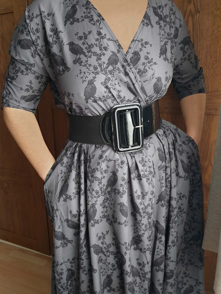 Gray dress with black ravens & blossoms pattern and black belt worn by a person against a wooden door background
